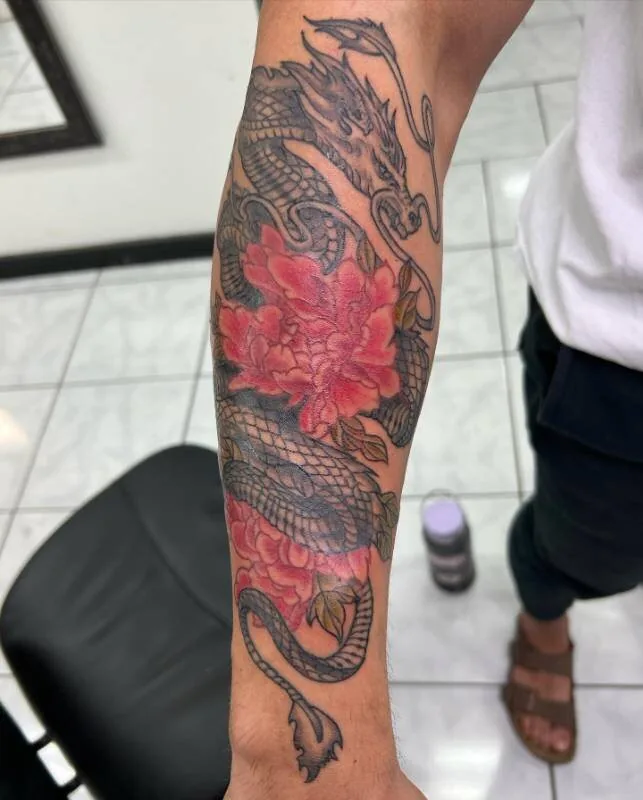 Black and grey Japanese dragon tattoo coiling through saturated red peonies down a forearm sleeve, by Christian "Tiki" Ramos at Ohana Tattoo Company, Waikiki, Hawaii.