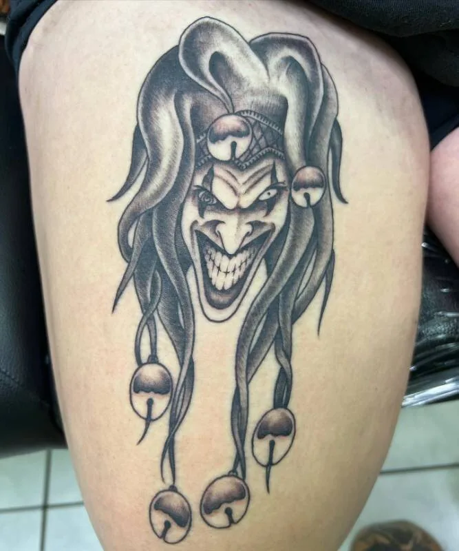 Black and grey tattoo of a sinister grinning court jester wearing a two-pointed cap with bells trailing down to the knee, on the thigh, by Christian "Tiki" Ramos at Ohana Tattoo Company, Waikiki, Hawaii.