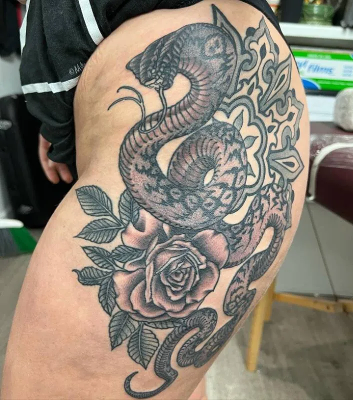 Black and grey tattoo of a cobra coiled over a rose with an ornamental Victorian scroll behind it, on the hip and thigh, by Christian "Tiki" Ramos at Ohana Tattoo Company, Waikiki, Hawaii.